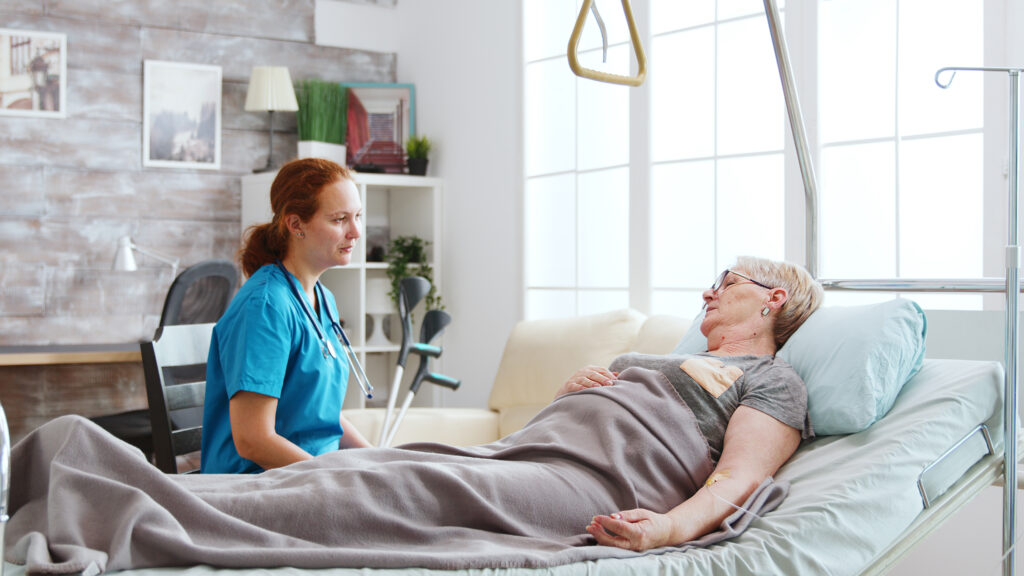 Nurse in retirement home talking with an old lady lying in hospital bed. Big windows with bright light are behind